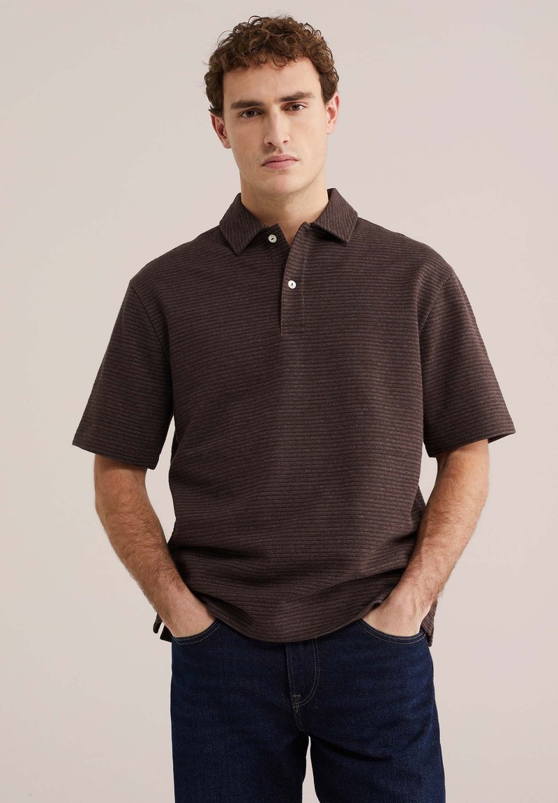 Homme aux cheveux bouclés portant un polo à manches courtes marron foncé et un jean bleu foncé, debout les mains dans les poches sur un fond beige.