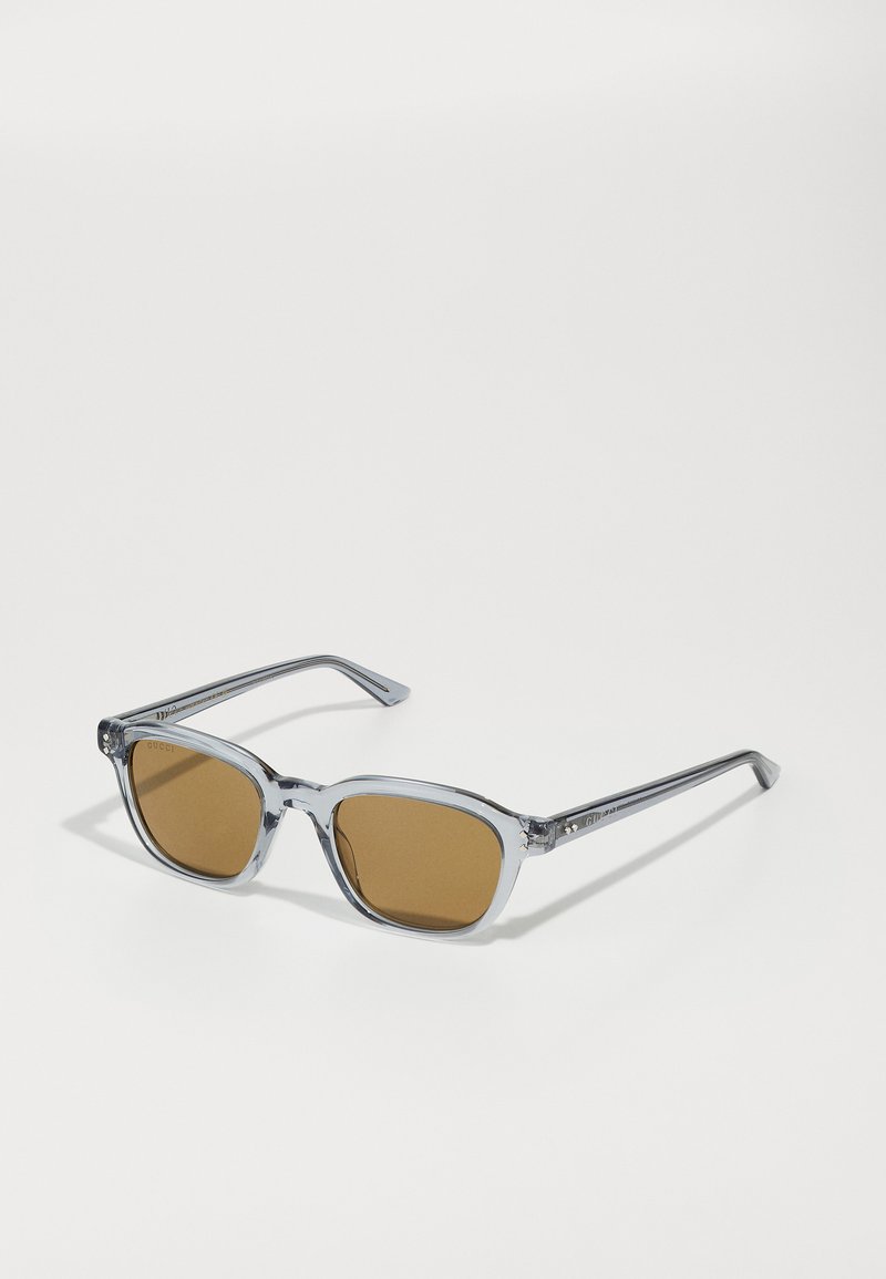 Sonnenbrille mit transparentem grauem Kunststoffrahmen, braunen Gläsern, quadratischer Form und Metallakzenten an den Bügeln. Einfaches Design, ohne Muster.