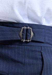 Pantalon bleu foncé à fines rayures avec une ceinture à boucle carrée argentée, porté avec une chemise blanche rentrée.