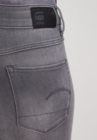 Jeans de mezclilla gris con una etiqueta de cuero negra en la cintura, detalles de costura sutiles y un bolsillo trasero con diseño de costura curvada.