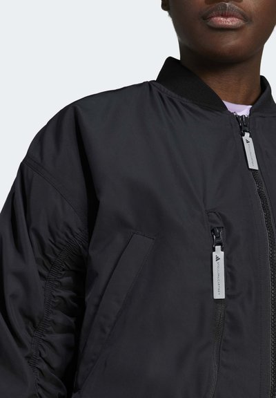 Közelkép egy személyről, aki fekete dzsekit visel, látható cipzárokkal és zsebekkel, látható a gallér és a felső mellkas területe.