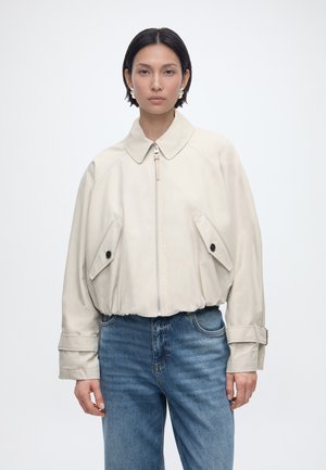 Ženska s kratkimi črnimi lasmi, oblečena v svetlo bež jakno na zadrgo z velikimi žepi s preklopom in modrimi kavbojkami, stoji na enobarvni podlagi.