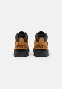 Adidași înalți din suede maro și sintetic negru, cu un logo proeminent „Timberland” pe călcâi. Prezintă șireturi rotunde și o talpă texturată.