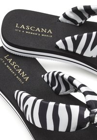 Schwarz-weiße Flip-Flops mit zebra-gestreiften Stoffriemen, strukturiertem Gummi-Fußbett und einer kontrastierenden weißen Sohle. Markenname in Gold aufgedruckt.