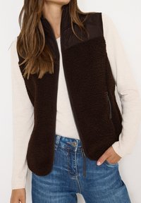 Chaleco de forro polar marrón con una sección superior acolchada, cierre de cremallera, bolsillos laterales y una textura suave, combinado con un suéter ligero y unos pantalones vaqueros.