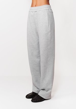 Osoba nosząca jasnoszare, luźne spodnie dresowe w połączeniu z czarnymi, marszczonymi butami wsuwanymi, pokazana od talii do stóp na białym tle.