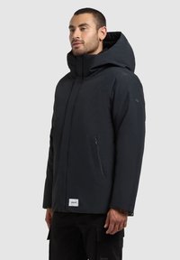 Černá izolační bunda s vysokým límcem, velkou kapucí a bočním kapsou na zip. Hladká textura a minimalistický design, obsahující malou nášivku s logem.