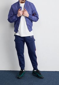 Man som bär en blå bomberjacka, vit t-shirt, marinblå joggingsbyxor med fickor för att lasta, och svarta-gröna sneakers stående på en blå matta.