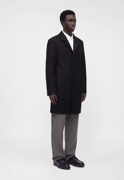 Jeune homme debout, portant un manteau noir boutonné, une chemise blanche, un pantalon gris et des chaussures noires, sur un fond clair uni.