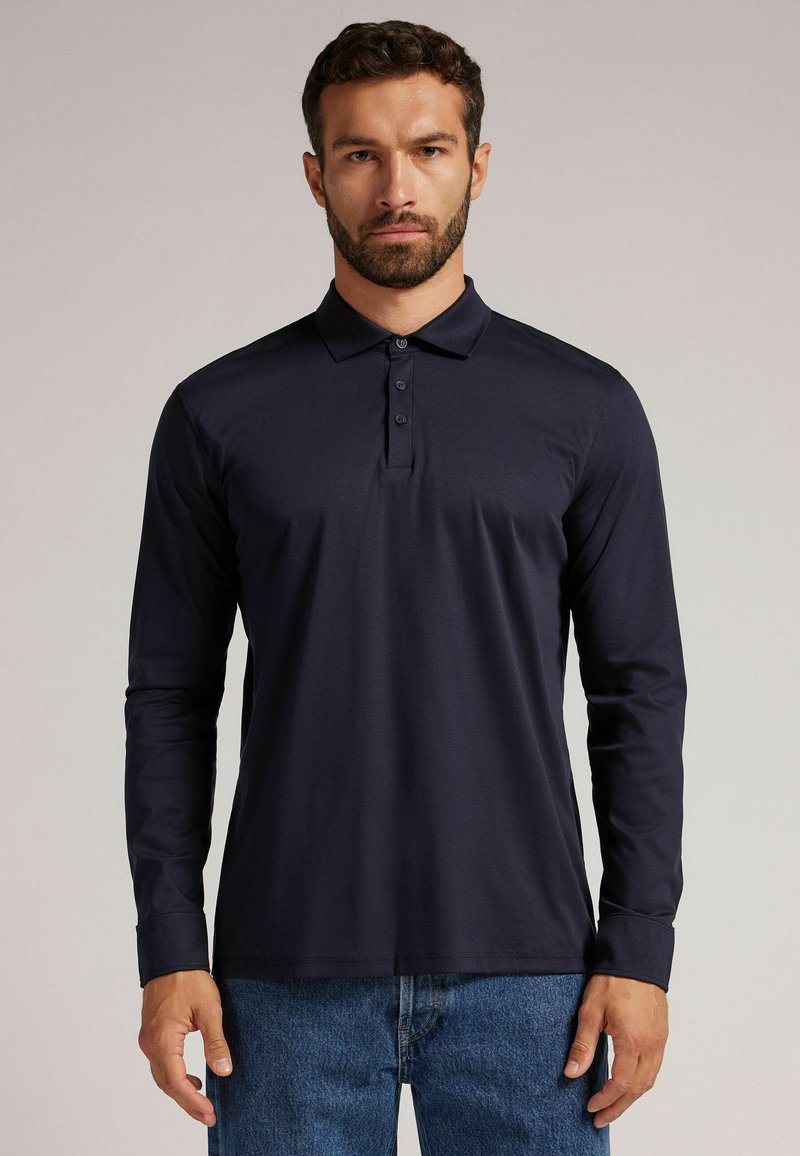 Långärmad polo i marinblått med en klassisk krage och fyrknäppt knappslå. Mjukt tyg med en slät textur, matchat med denimjeans.