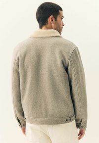 Grå uldjakke med en faux shearling krave, der har knaplukning foran, sideknapper og en tekstureret overflade. Simpelt og struktureret design.