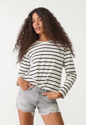 Gestreiftes Langarmoberteil in Off-White und Schwarz mit Rundhalsausschnitt, kombiniert mit hellgrauen, zerrissenen Denim-Shorts mit ausgefransten Säumen.