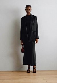 Svart skreddersydd blazer over en tettsittende svart kjole, kombinert med svarte stiletter med stropper og en liten rød håndveske. Glatt stoff, minimalistisk design.