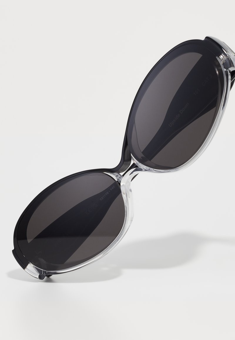 Occhiali da sole oversize neri con montatura trasparente, lenti scure sfumate e una texture liscia e lucida. Presentano bordi arrotondati e senza loghi visibili.