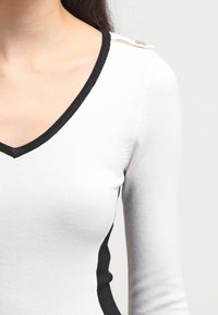 Pull à col en V blanc avec bordures et accents noirs. Il présente des manches longues et un détail décoratif avec bouton sur l'épaule. Tissu texturé.