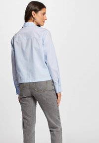 Lichtblauwe cropped blouse met lange mouwen, knoopsluiting en een gladde textuur, gecombineerd met grijze jeans met achterzakken en stiksels.
