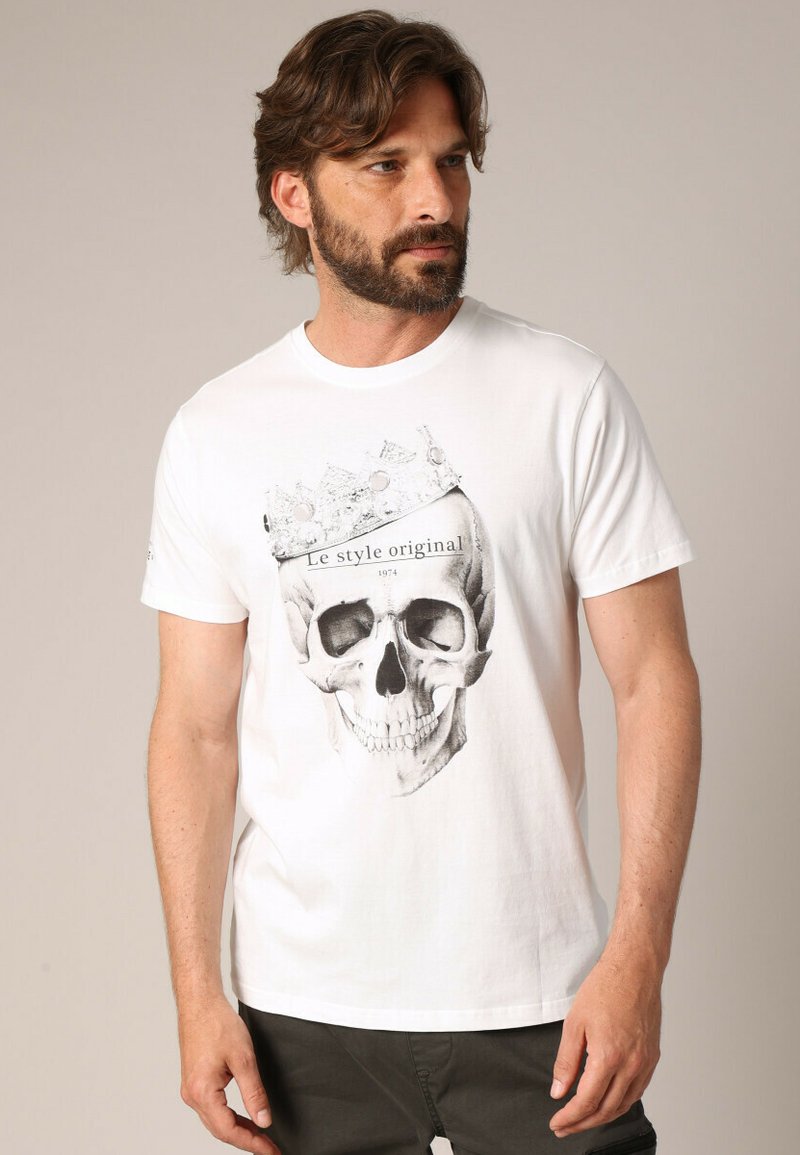 T-shirt en coton blanc avec un graphique d'un crâne portant une couronne, impression noire et texte "Le style original 1974." Manches courtes.