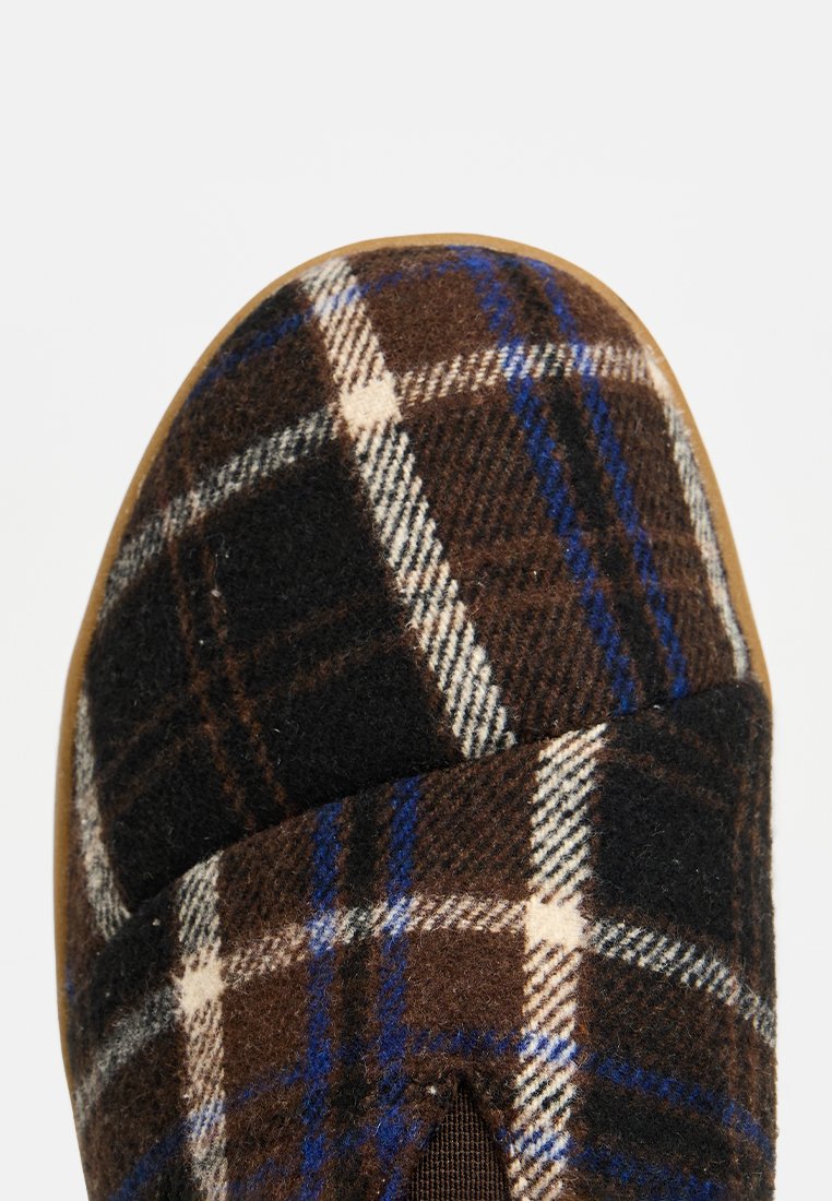 Zapatilla de cuadros marrón con textura de lana, líneas azules y blancas en un patrón de cuadros, con punta redondeada y suela de goma.