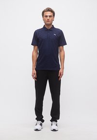 Modèle masculin debout portant un polo bleu marine, un pantalon noir et des baskets blanches sur un fond blanc uni.