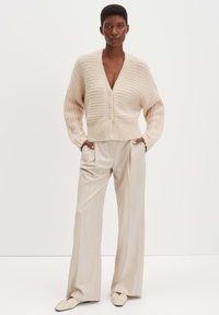 Helle beige Strickjacke mit V-Ausschnitt, strukturierten Streifen, fünf Knöpfen und langen Ärmeln, kombiniert mit cremefarbenen, weit geschnittenen Hosen und flachen Schuhen.