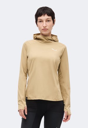 Γυναίκα που φοράει μπεζ μακρυμάνικη μπλούζα με κουκούλα και μικρό λευκό λογότυπο στο στήθος και μαύρο παντελόνι, στέκεται μπροστά σε απλό φόντο.