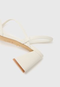 Sandale blanche à talons hauts avec des brides croisées, fabriquée à partir d'un matériau lisse. Talon carré avec un accent brun clair à la base.