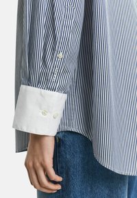 Chemise rayée bleue et blanche avec un poignet blanc, dotée de boutons et d'un subtil détail de logo sur la manche. Texture en coton évidente.