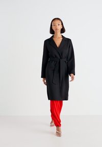 Black long coat with a belt, featuring a collar and side pockets, paired with red trousers and silver sandals, set against a light background.