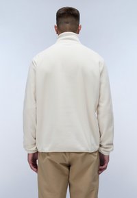 Kræmfarvet fleecepullover med høj krave, lange ærmer og lige kant. Blød, struktureret overflade. Båret med beige bukser.