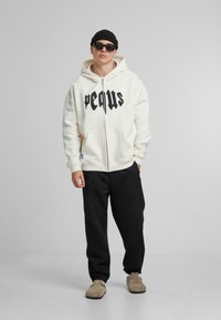 Crème hoodie met ritssluiting en zwarte tekst, met voorzakken, gecombineerd met zwarte relaxte broek en beige schoenen. Zwarte beanie en zonnebril gedragen.