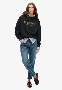Felpa nera con il logo "SuperDry", camicia a righe blu e bianche sotto, jeans azzurri chiari e mocassini scuri. Vestibilità rilassata e design casual.