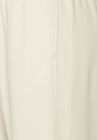 Beige sweatpants lavet af blødt stof, med et hvidt broderet logo på siden. Indeholder sidelomme og elastisk talje.