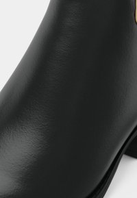 Črni usnjeni gležnjar z gladko teksturo, okrogel sprednji del in subtilen zlati poudarek na strani.