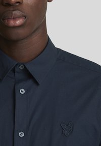 Camicia in cotone blu navy con colletto a punta, dotata di bottoni coordinati e un piccolo logo di farfalla ricamato sul lato sinistro del petto.