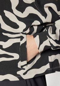 Sort og beige sweatshirt med et abstrakt bølgemønster, tætsiddende manchetter og en sidelomme synlig nær taljen.