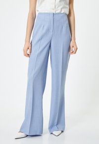 Pantaloni ampi di un blu chiaro con una vestibilità sartoriale, realizzati in tessuto morbido. Presentano una vita alta e pieghe frontali, abbinati a scarpe bianche a punta.