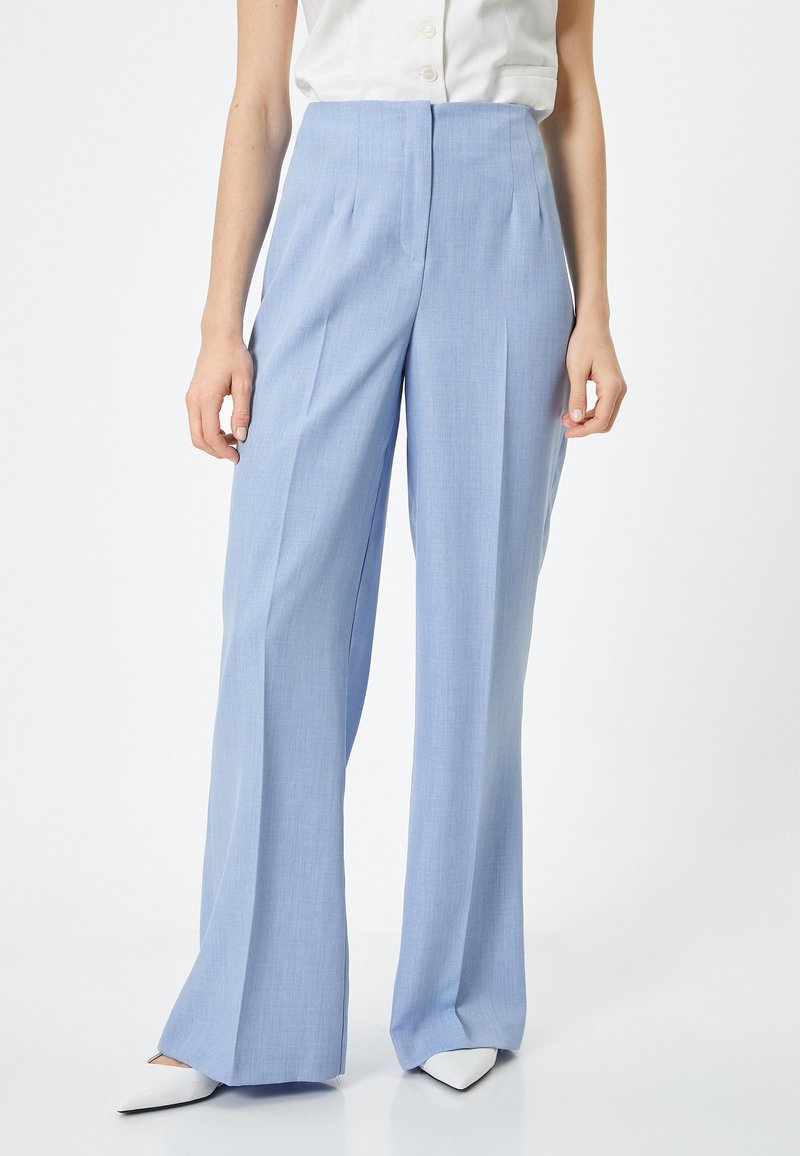 Pantaloni ampi di un blu chiaro con una vestibilità sartoriale, realizzati in tessuto morbido. Presentano una vita alta e pieghe frontali, abbinati a scarpe bianche a punta.