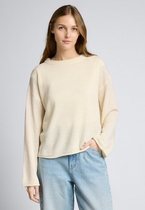 Jeune femme aux cheveux longs portant un pull en maille crème et un jean bleu clair, posant avec une main dans la poche devant un fond gris.