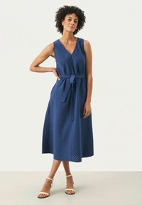 Marineblauwe, mouwloze jurk met een V-hals, textuurstof en een strikceintuur in de taille. Valt onder de knie met een uitlopende rok.