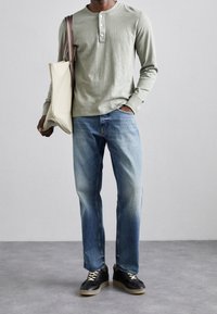 Ljusgrön långärmad henleytröja med tre knappar, blå jeans och svarta sneakers med beige sulor, bärande en beige toteväska.