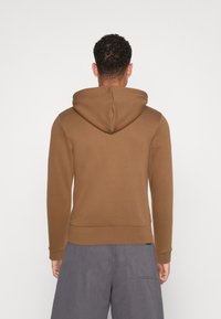 Sweat à capuche marron avec une grande capuche, des poignets et un ourlet côtelés. Le matériau semble doux avec une texture lisse. Porté avec un short gris.