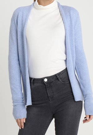 Femme portant un cardigan bleu clair ouvert sur un col roulé blanc rentré dans un jean noir taille haute, debout, mains détendues.