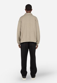 Bež srajca z dolgimi rokavi in teksturo v obliki vafljev s ovratnikom, kombinirana z ohlapnimi črnimi hlačami in supergami, prikazana s hrbtne strani.
