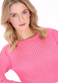 Różowy sweter w paski z dzianiny o prążkowanej fakturze i okrągłym dekolcie; włosy modelki ułożone w luźne fale.