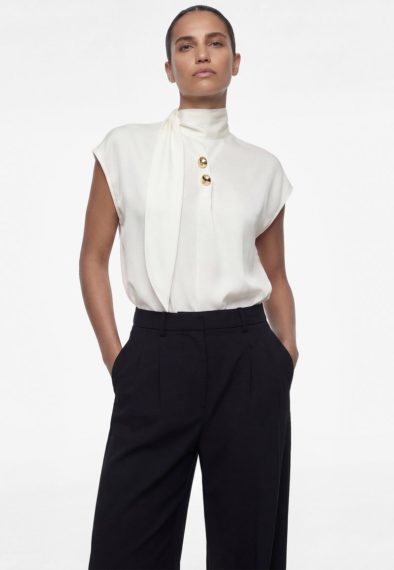 Mujer con blusa blanca sin mangas de cuello alto y botones dorados, combinada con pantalones negros sueltos de talle alto, con las manos en los bolsillos.