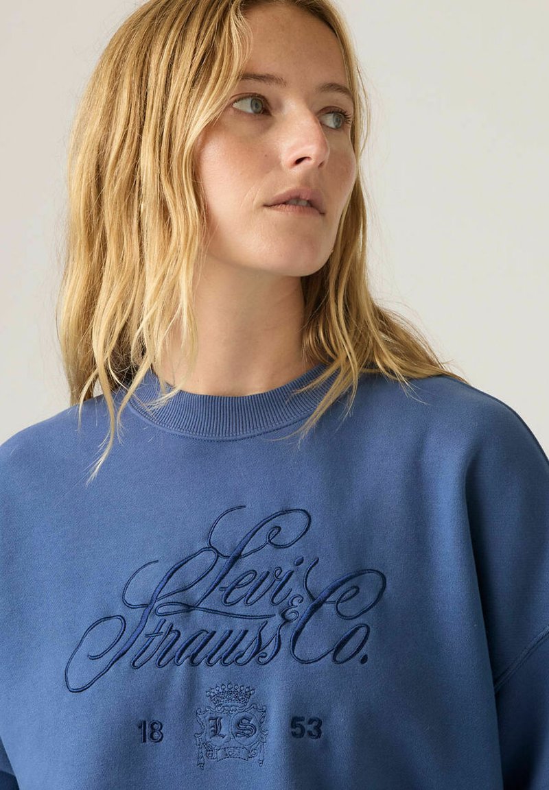 Femme blonde regardant vers la droite, portant un sweat-shirt bleu Levi's Strauss & Co. avec un texte brodé et un blason sur la poitrine.