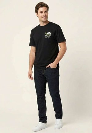 Hombre sonriente de pie, lleva una camiseta negra con diseño gráfico, vaqueros oscuros y zapatillas blancas, con una mano en el bolsillo contra un fondo sencillo.