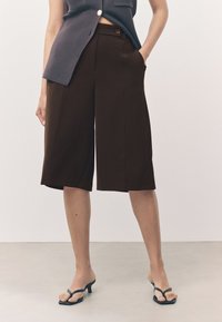 Pantalones marrones de pierna ancha y corte cropped con bolsillos, textura suave y un corte ligeramente entallado, combinados con sandalias de tacón negras con tiras.