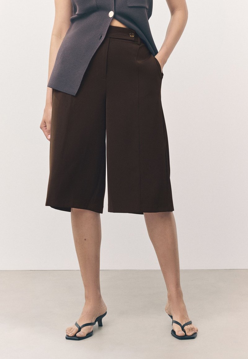 Pantalones marrones de pierna ancha y corte cropped con bolsillos, textura suave y un corte ligeramente entallado, combinados con sandalias de tacón negras con tiras.