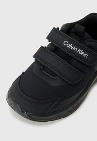 Chaussure de sport noire en tissu et caoutchouc, dotée de deux bandes Velcro, d'une semelle texturée et d'un logo "Calvin Klein" blanc.